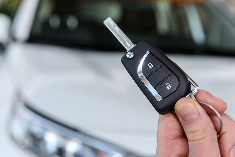 A hand holding a car key fob with lock and unlock buttons, with a blurred white car in the background.