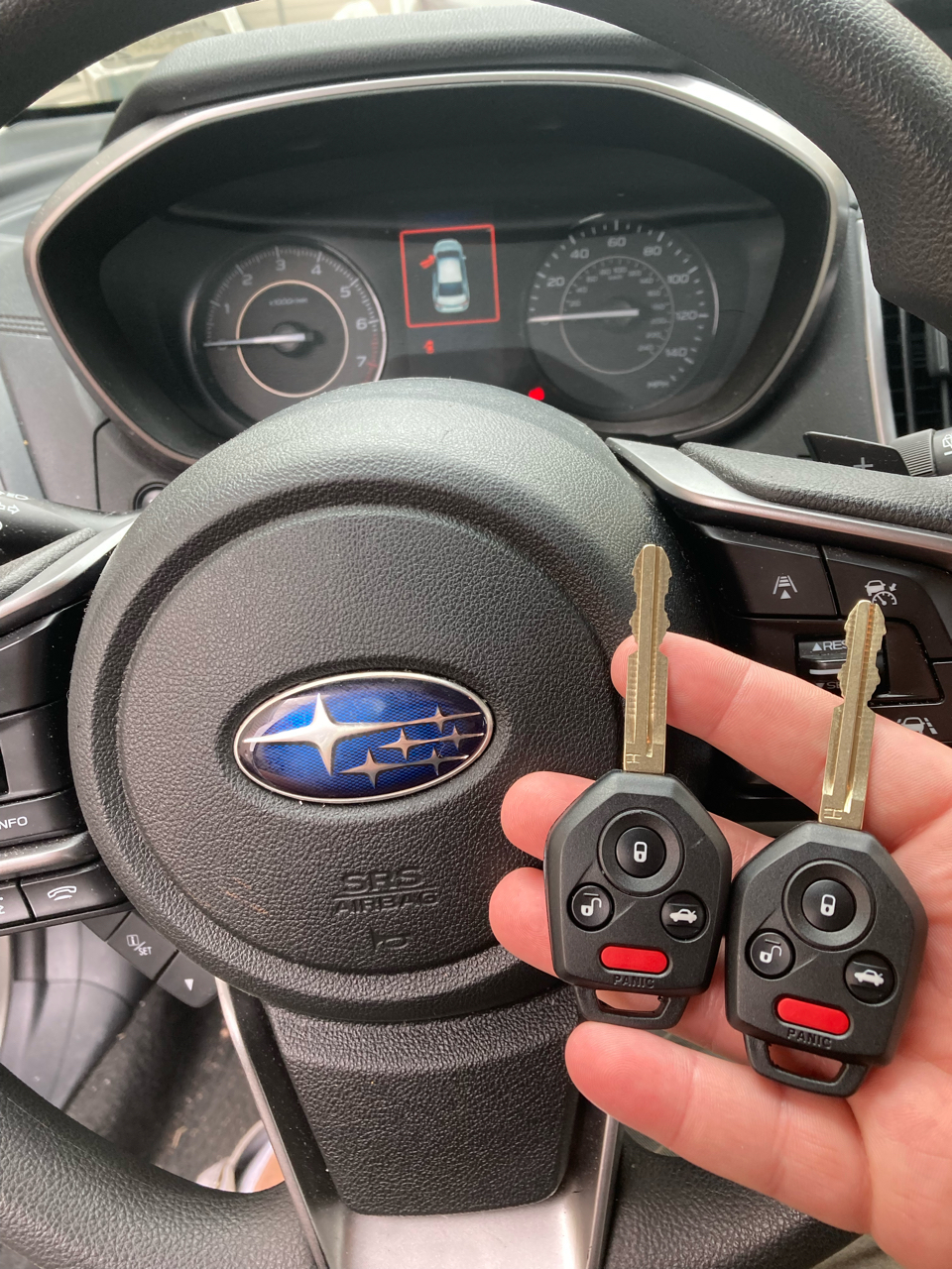 A hand holds two Subaru car keys with remote buttons in front of a Subaru steering wheel and dashboard.
