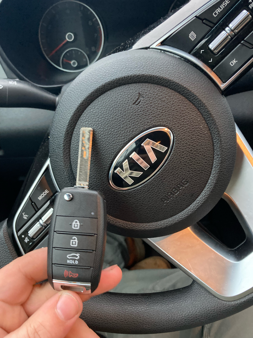 A hand holds a Kia car key fob with an extended key blade in front of a Kia steering wheel and dashboard.