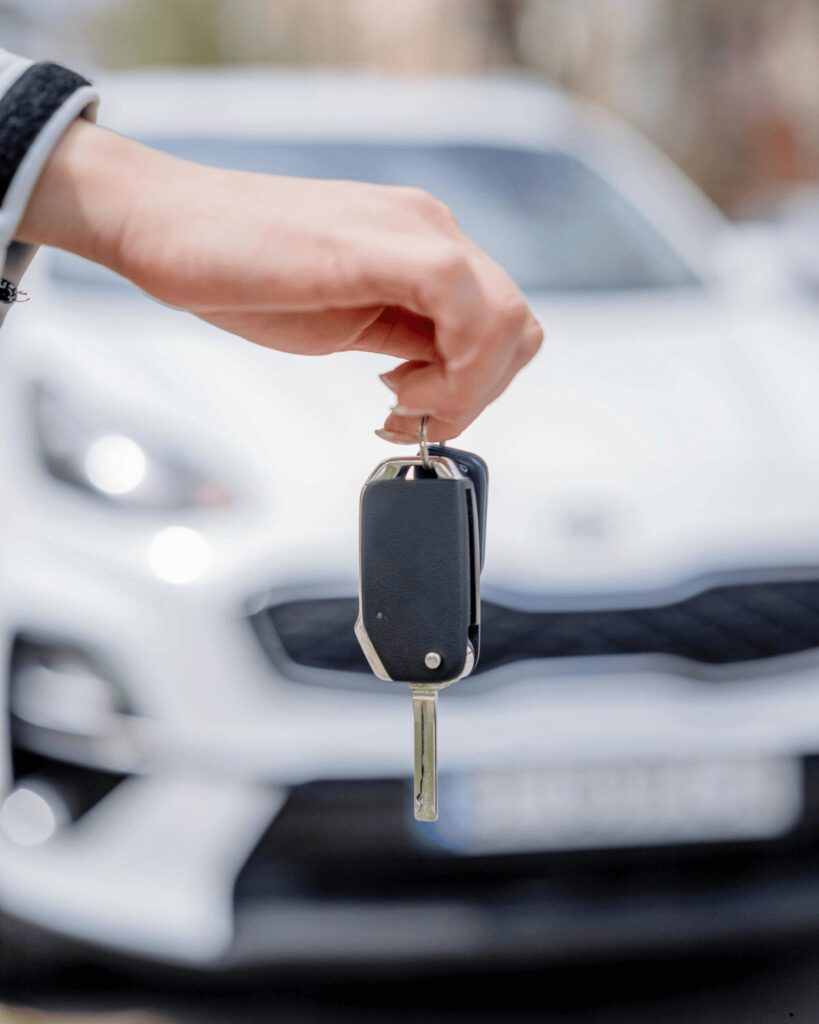 A hand holding a car key fob with a blurred white car in the background, perfect for illustrating Car Key Replacement services.