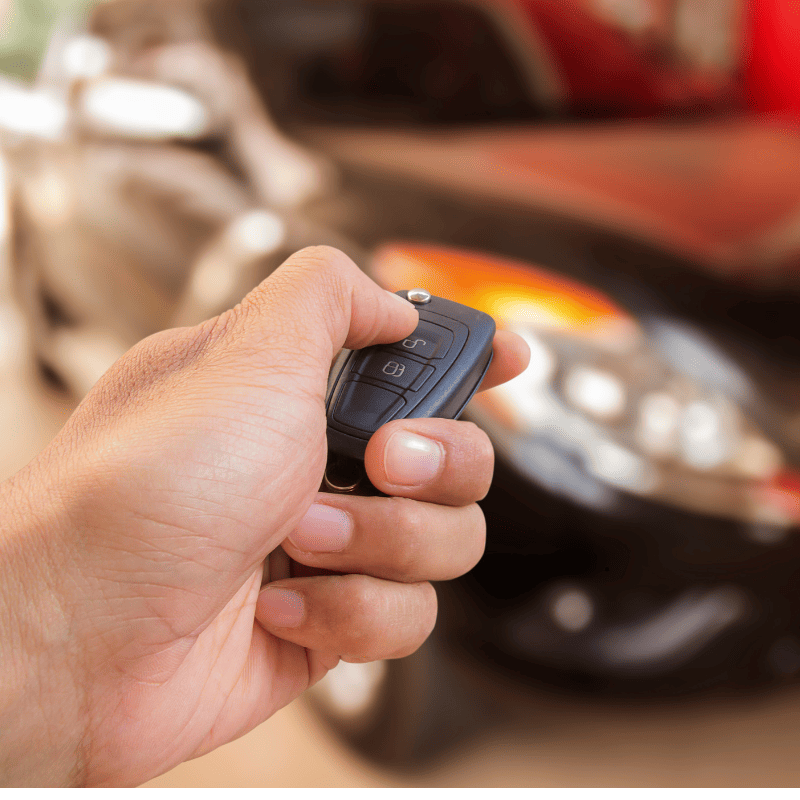 A hand holding a car key fob in front of a blurred black car, highlighting convenient Car Key Replacement solutions.