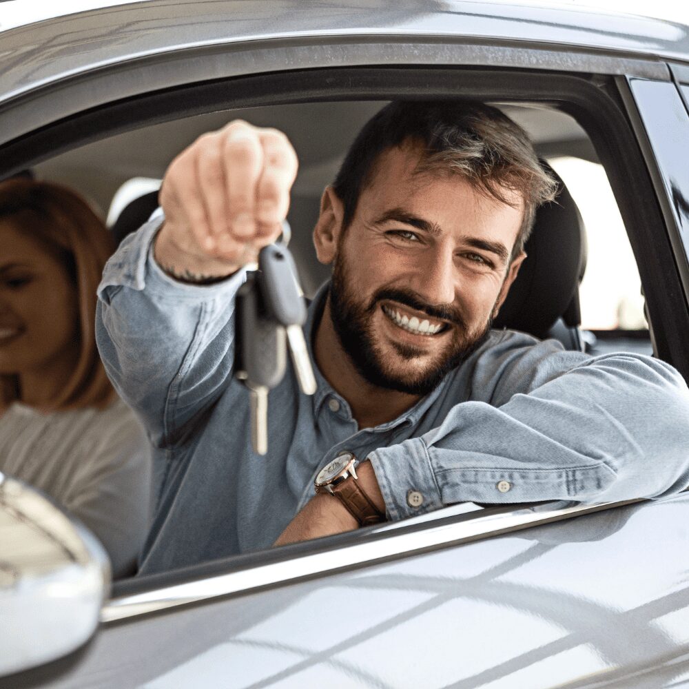 A man sitting in the driver’s seat of a car smiles and holds out a set of car keys, showing off his recent Car Key Replacement, while a woman sits in the passenger seat. A man sitting in the driver's seat of a car smiles and holds out a set of car keys, showing off his recent Car Key Replacement, while a woman sits in the passenger seat.