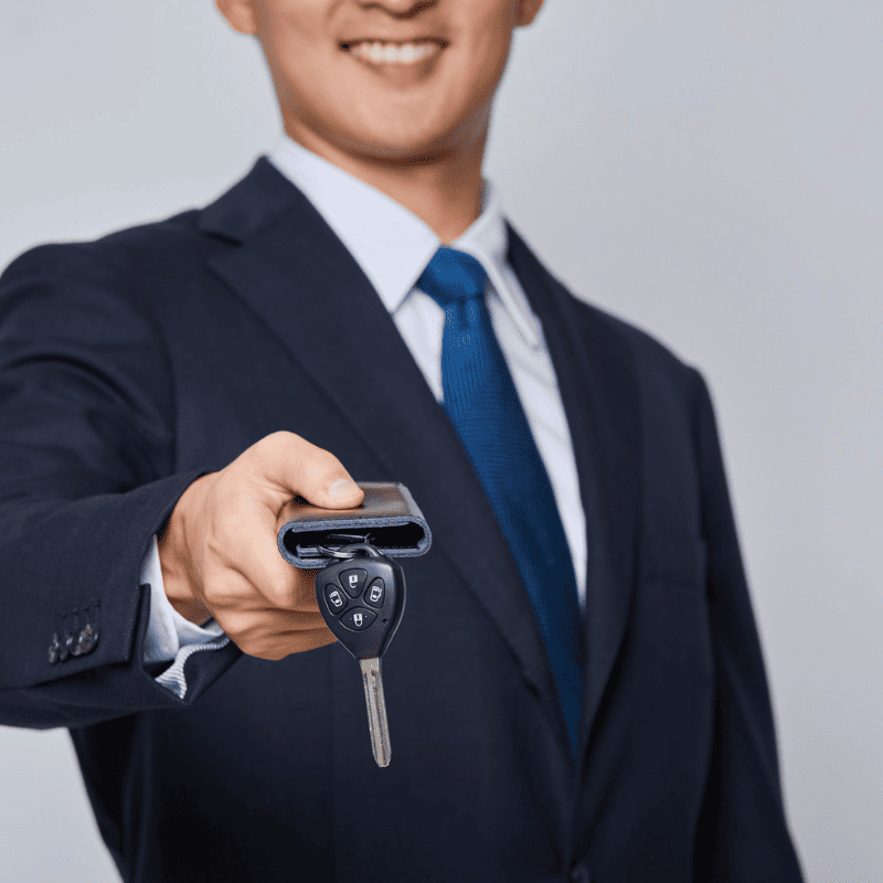 A man in a suit and tie holds out a car key and remote towards the camera, smiling slightly, representing a trusted locksmith service in North San Bernardino.