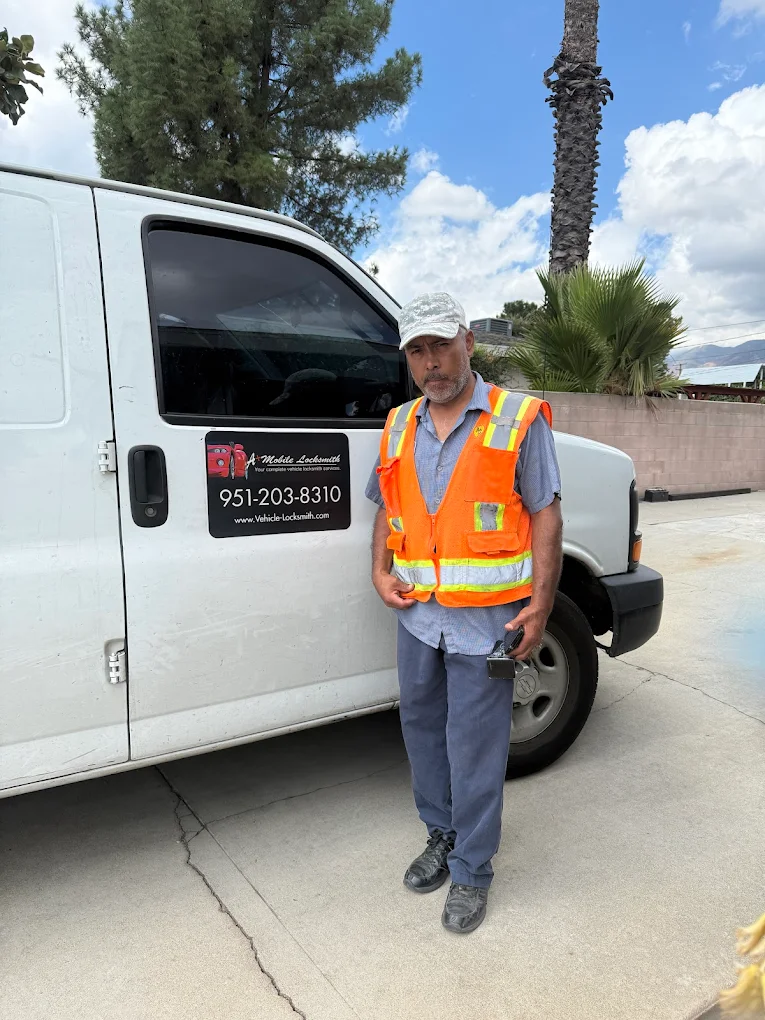 A man in a high-visibility safety vest stands beside a white work van with a company sign and phone number on the side. He is holding a cellphone and standing on a driveway.