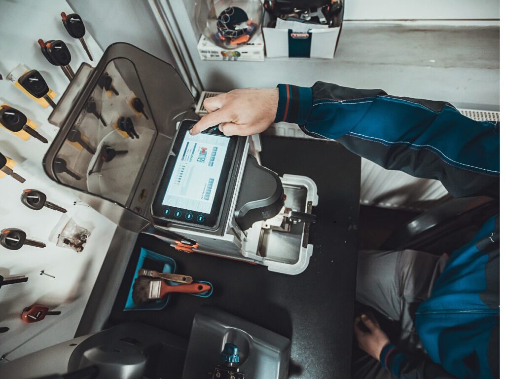 A Locksmith Banning expert operates a digital key cutting machine at a workstation, surrounded by tools hanging on the wall and specialized equipment on the desk.