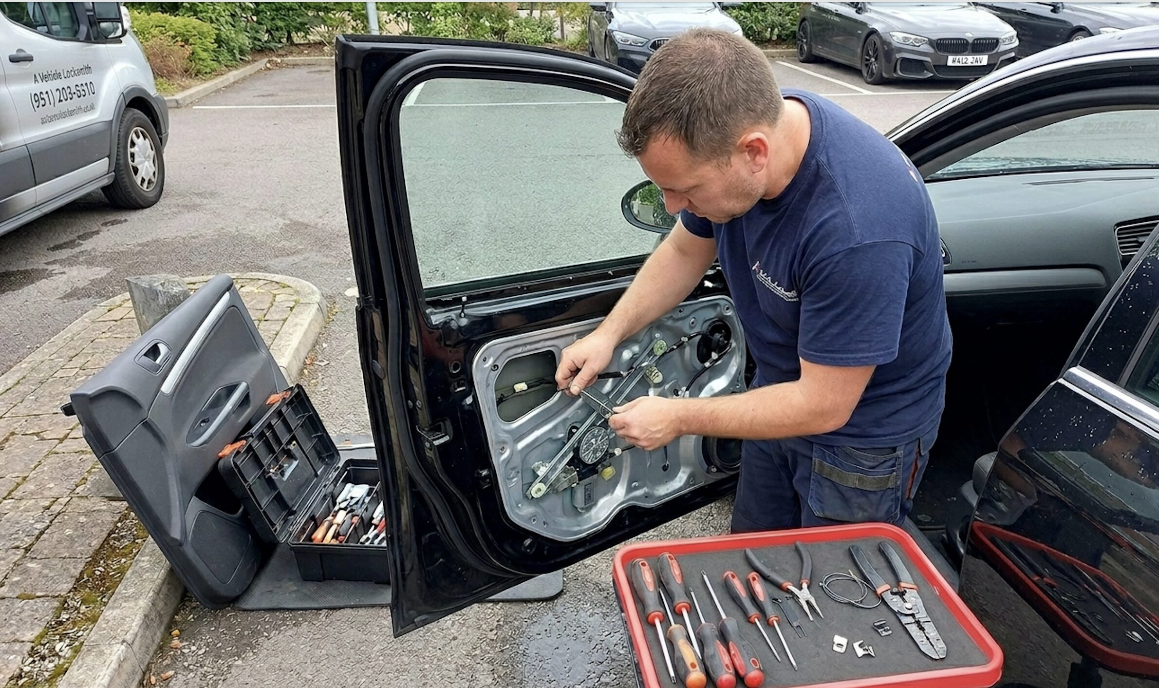 A locksmith repairs the inside of a car door with the panel removed, using tools from a toolbox and tray beside him in a Highland parking lot.
