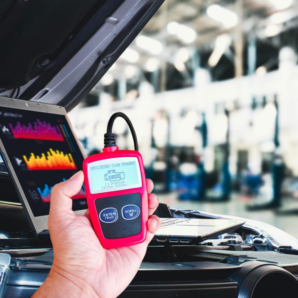 Technician holding an OBD2 code reader in front of an open car hood while a laptop displays diagnostic and car key programming San Bernardino data inside a professional auto repair shop.